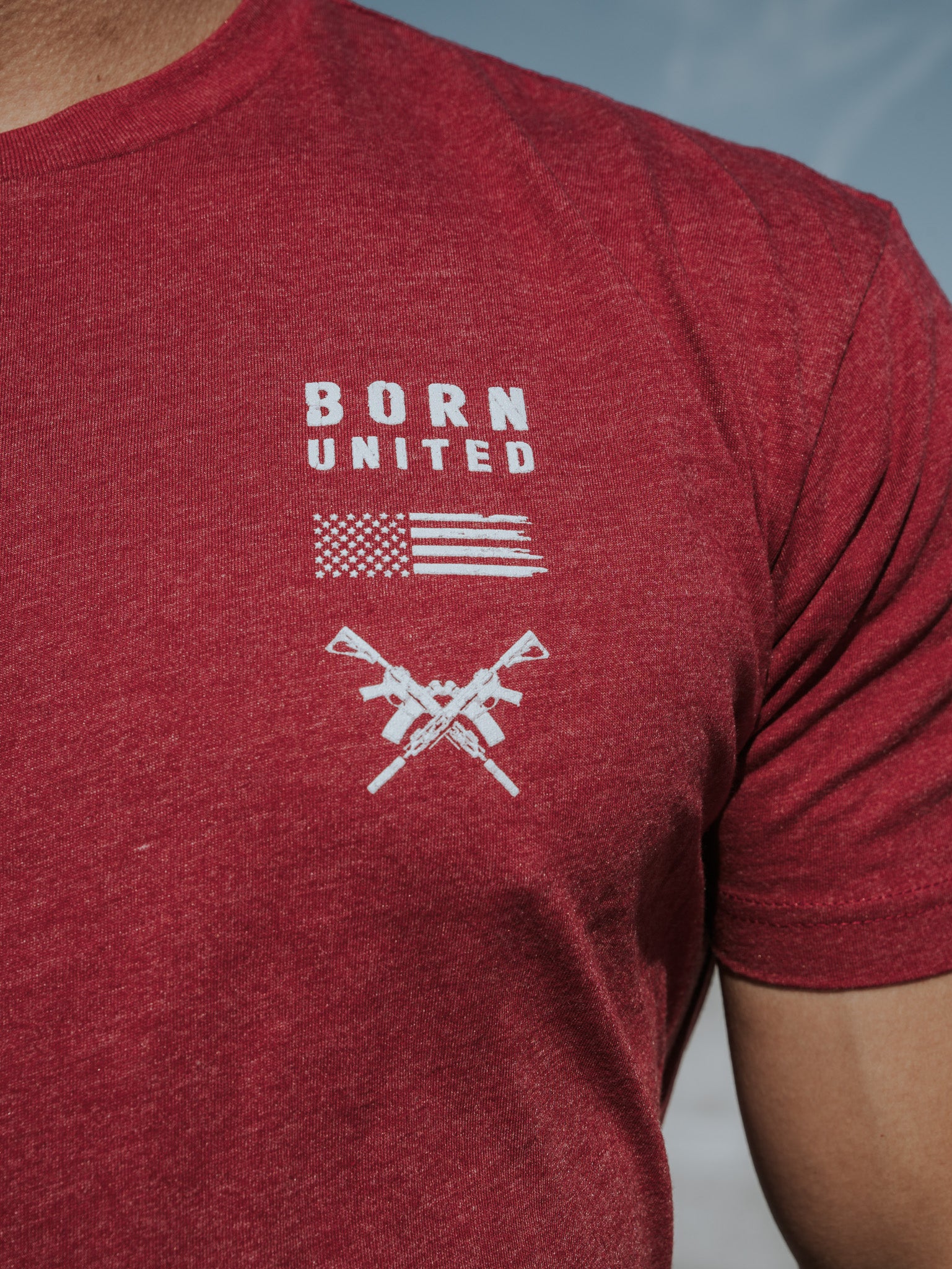 A person wears the R.E.D. Friday T-shirt by Born United Private Label, featuring BORN UNITED text above a stylized American flag and crossed rifles, celebrating patriotism and honoring brave soldiers against a clear blue sky backdrop.