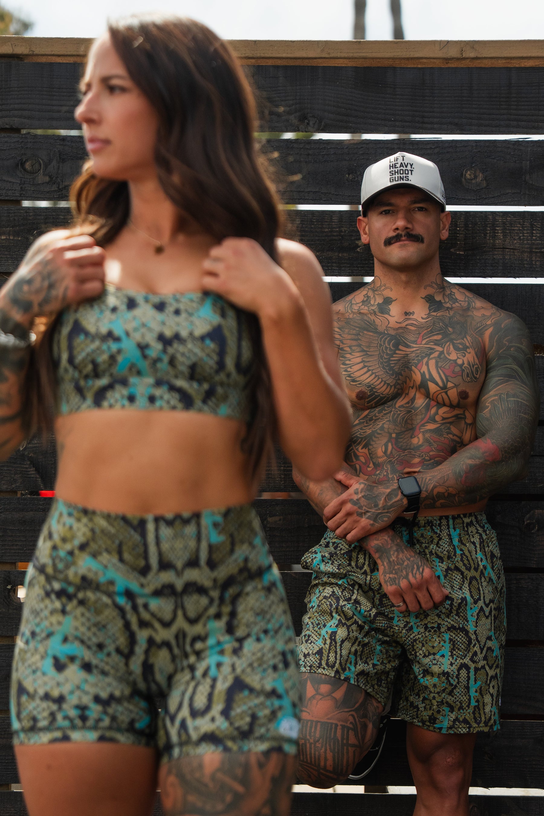 A woman wears Tango Charlie Apparel’s Lethal Venom Women’s High-Waisted Seamless Front Compression Training Shorts, her snakeskin pattern shorts in focus as she stands in the foreground; a tattooed man in matching shorts is by a dark wooden fence behind her.