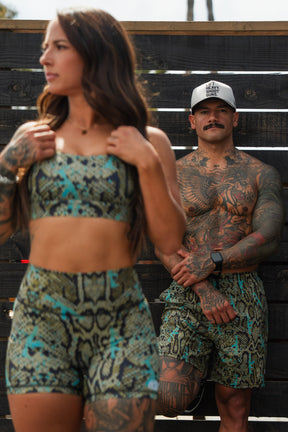 A woman wears Tango Charlie Apparel’s Lethal Venom Women’s High-Waisted Seamless Front Compression Training Shorts, her snakeskin pattern shorts in focus as she stands in the foreground; a tattooed man in matching shorts is by a dark wooden fence behind her.