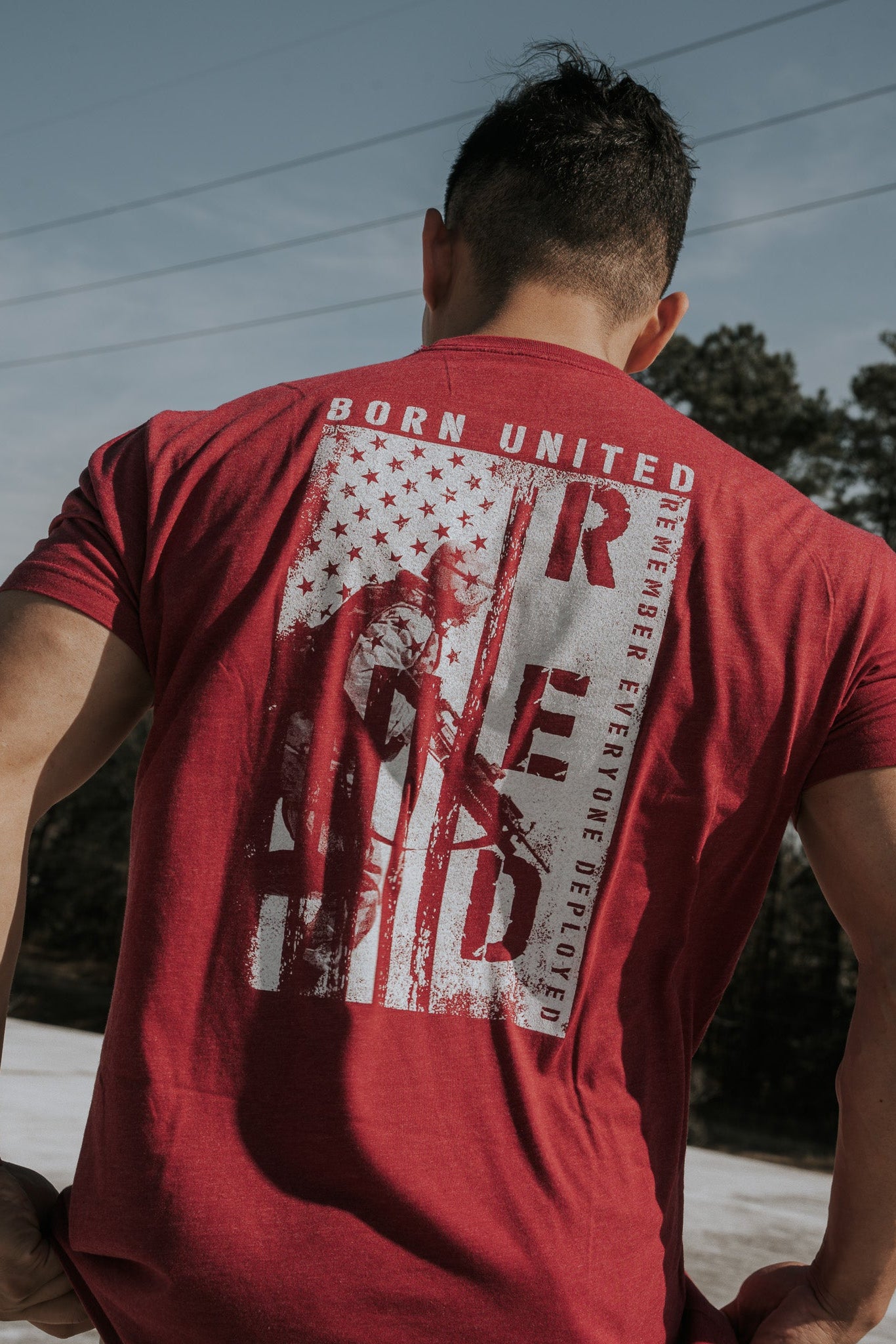 Someone outdoors wears a R.E.D. Friday shirt by Born United Private Label, featuring military-style graphics with BORN UNITED and R.E.D. Remember Everyone Deployed, honoring brave soldiers with a stylized flag and soldier image, celebrating the spirit of patriotism and freedom.