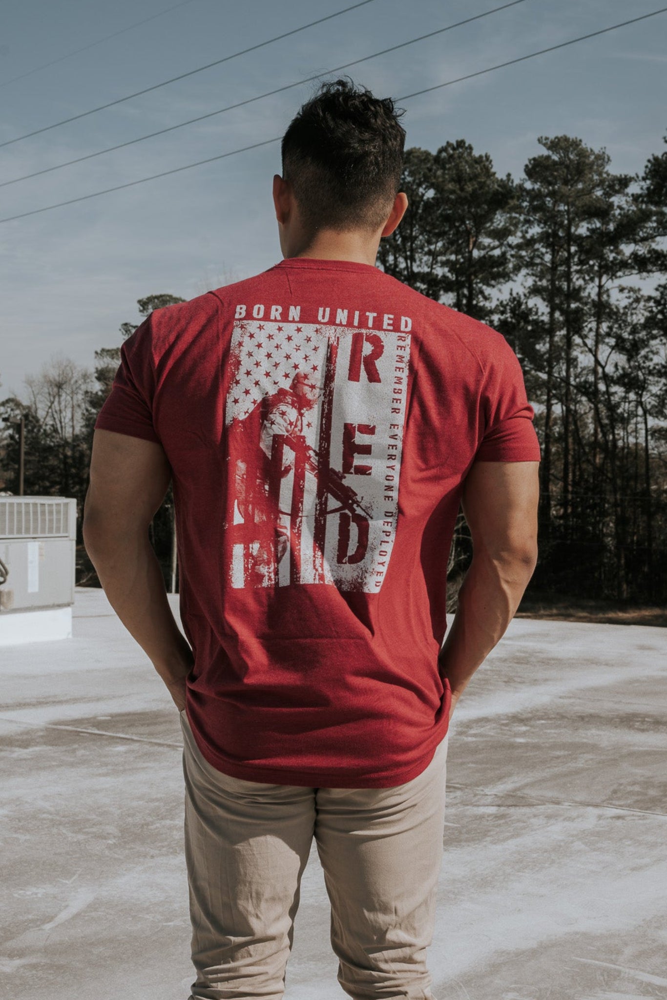 A man stands on a rooftop wearing a R.E.D. Friday shirt by Born United Private Label, facing away under a clear sky surrounded by trees, seemingly contemplating the freedoms protected by brave soldiers as power lines stretch across the horizon.