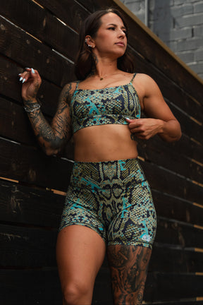 A woman with long brown hair and tattoos poses confidently against a dark wooden wall, wearing Tango Charlie Apparel’s Lethal Venom Women’s High-Waisted Seamless Front Compression Training Shorts—stylish and breathable for any workout.