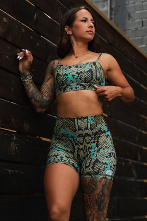 A tattooed woman poses confidently against a dark wooden wall, wearing the Straight Strap Sports Bra | Lethal Venom by Tango Charlie Apparel—a medium support, moisture-wicking set in green, blue, and yellow tones.