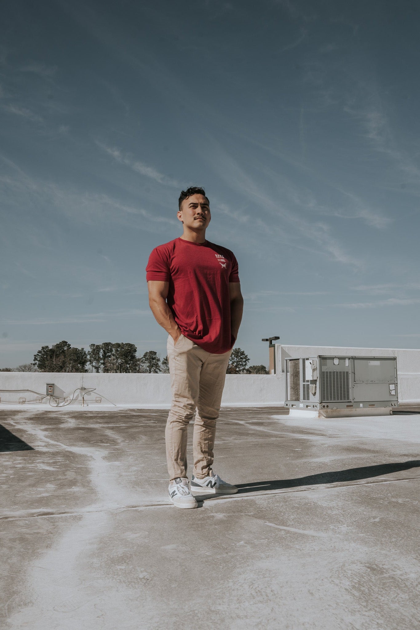 A man in a Born United Private Label R.E.D. Friday T-shirt and beige pants stands on a concrete rooftop, hands in pockets under a clear blue sky. Air conditioning units stand like sentinels behind him, embodying the quiet patriotism reflected in everyday life.