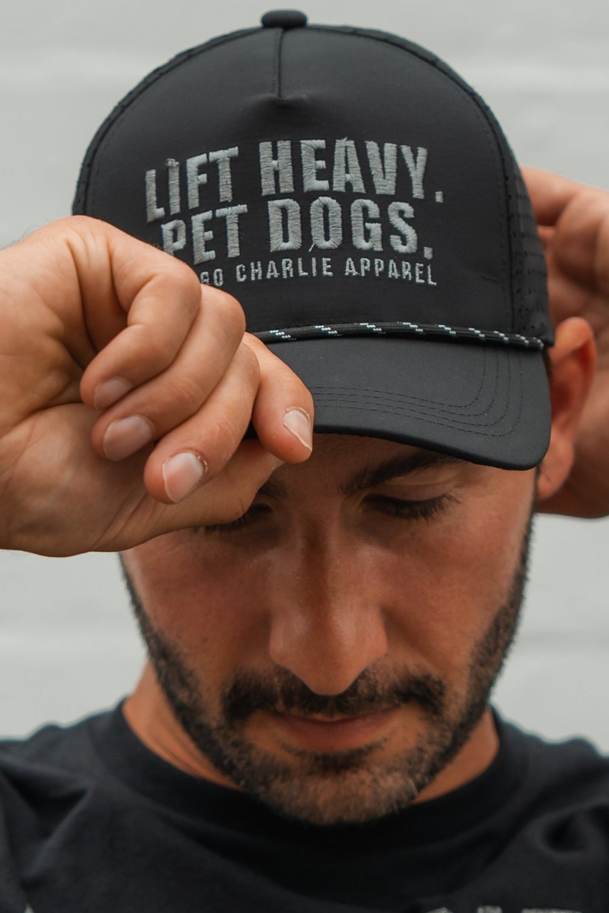 A bearded man looks down while adjusting his black Lift Heavy. Pet Dogs Snapback by Tango Charlie Apparel—perfect for lifting and dog lovers.
