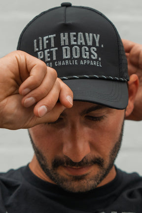 A bearded man looks down while adjusting his black Lift Heavy. Pet Dogs Snapback by Tango Charlie Apparel—perfect for lifting and dog lovers.