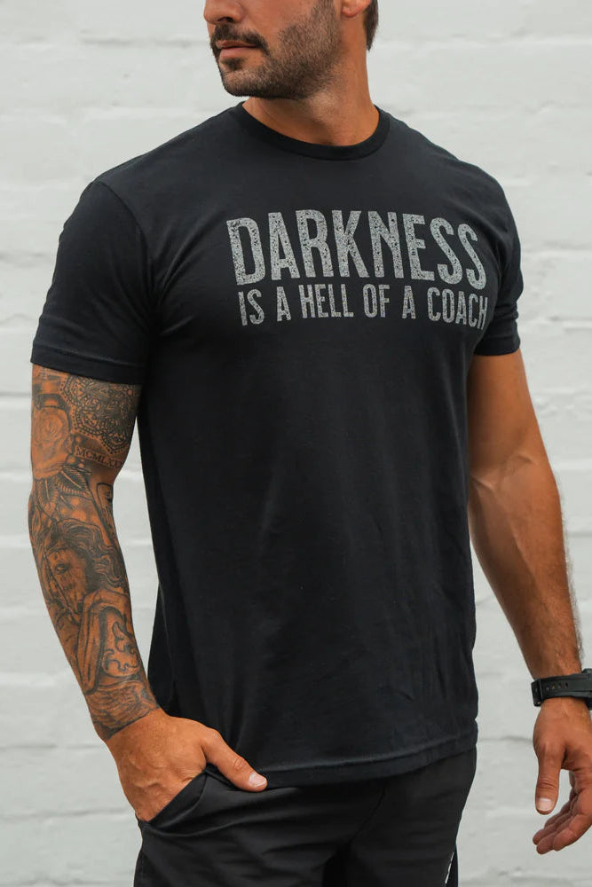 A tattooed man stands against a light wall, looking to the side with one hand in his pocket, wearing the Tango Charlie Apparel Darkness Is A Hell Of A Coach T-Shirt in black, made from perfect stretch ring-spun cotton.