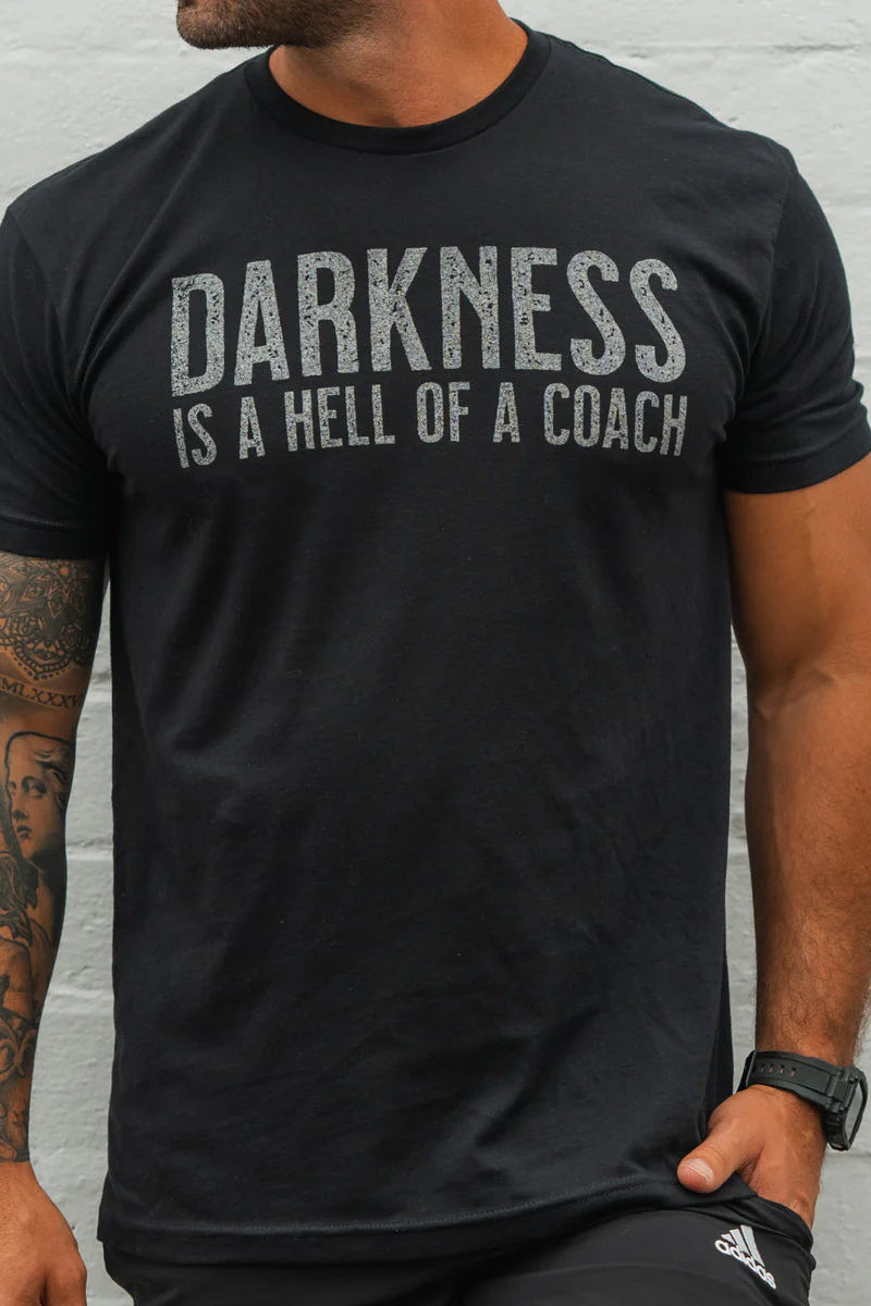 A man in the Darkness Is A Hell Of A Coach T-Shirt - Black by Tango Charlie Apparel, featuring bold white text, shows off his tattooed arm and watch against a light gray wall—embracing comfort and perfect stretch.
