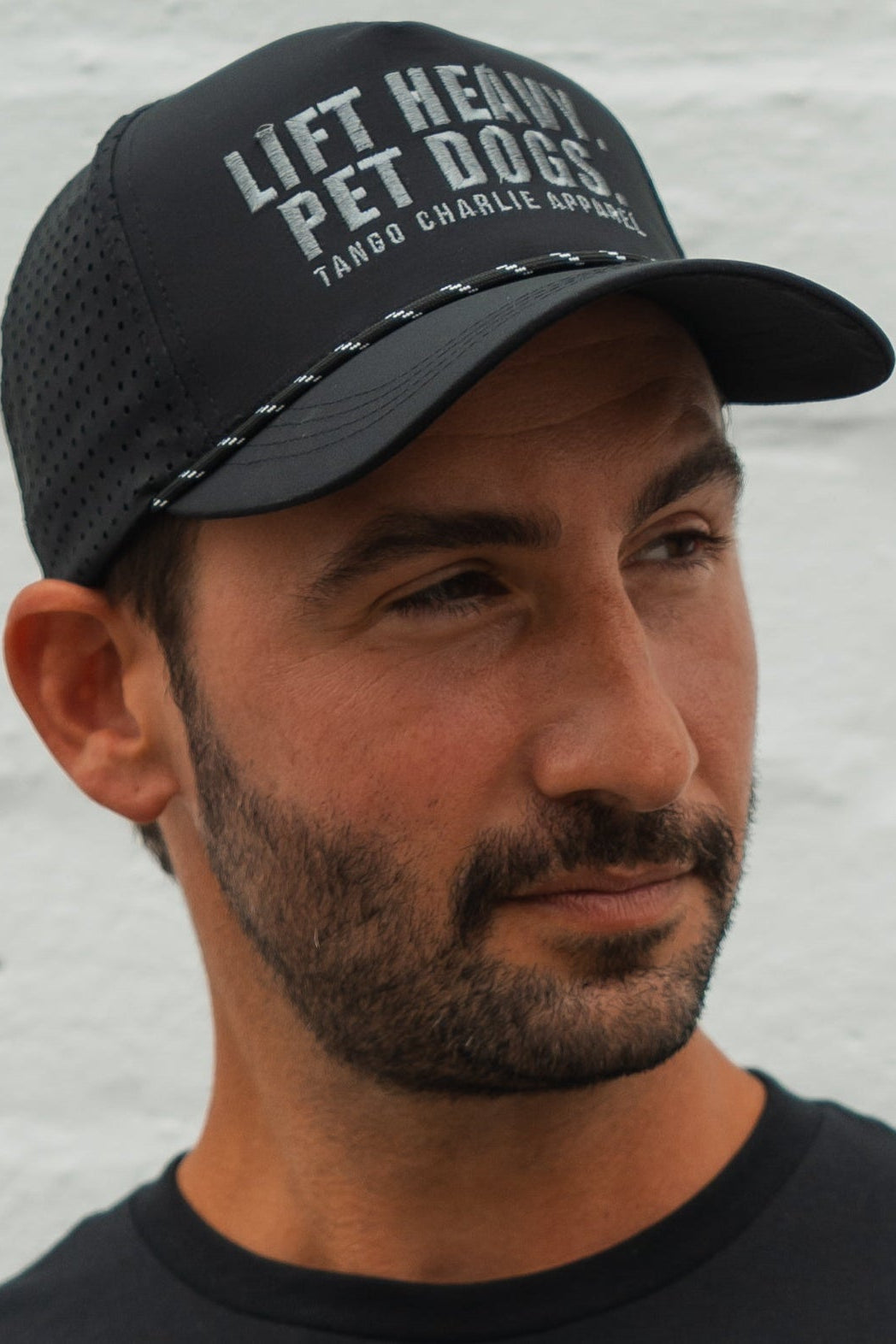 A bearded man wears the Tango Charlie Apparel Lift Heavy. Pet Dogs Snapback, a black, water-resistant hat with the slogan on front, as he looks to the side against a light background—ideal for lifting and dog lovers.