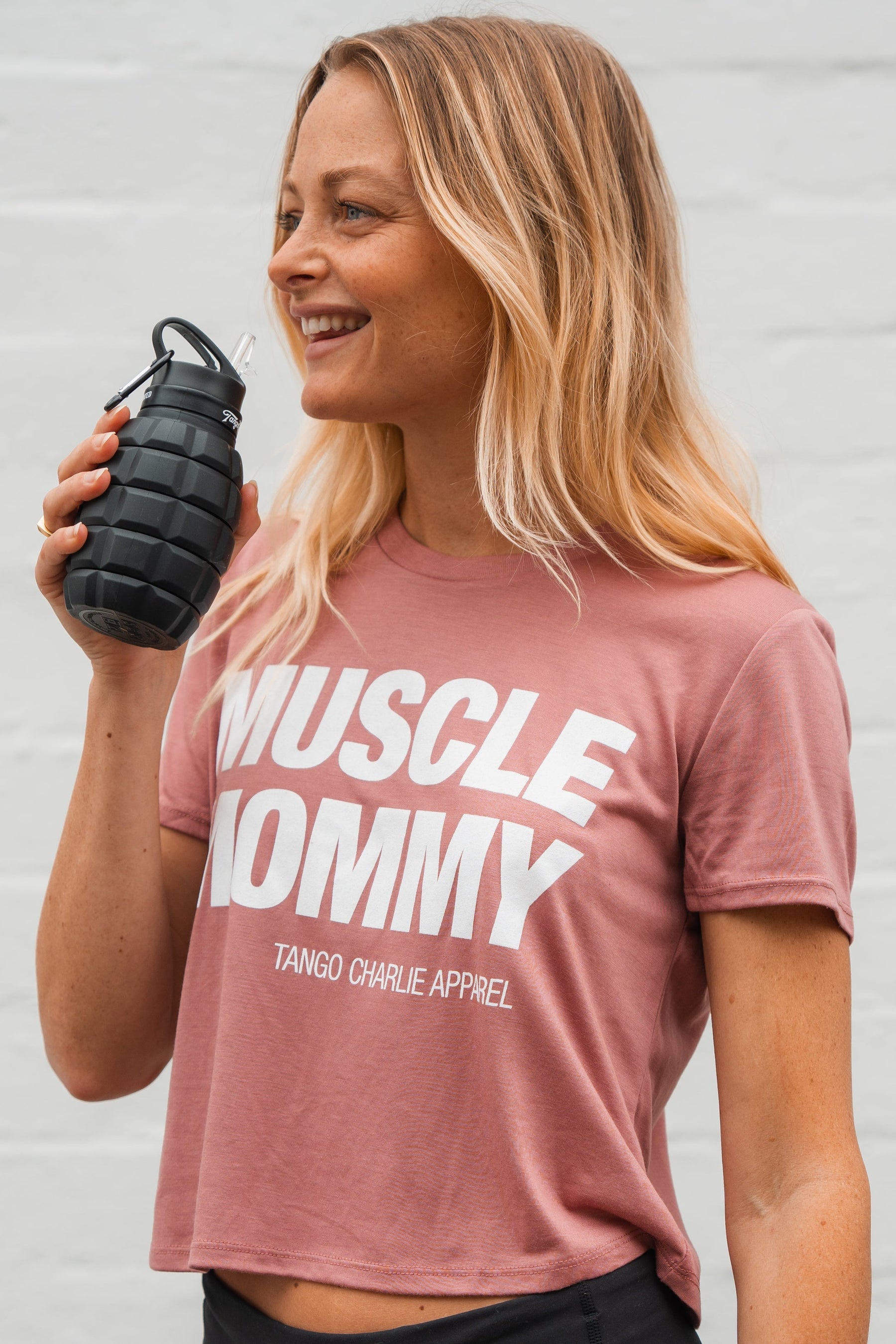 A fit, smiling woman with blonde hair wears the Muscle Mommy - Crop Tee by Tango Charlie Apparel and holds a black grenade-shaped water bottle—a true muscle mommy who can deadlift 225 lbs.