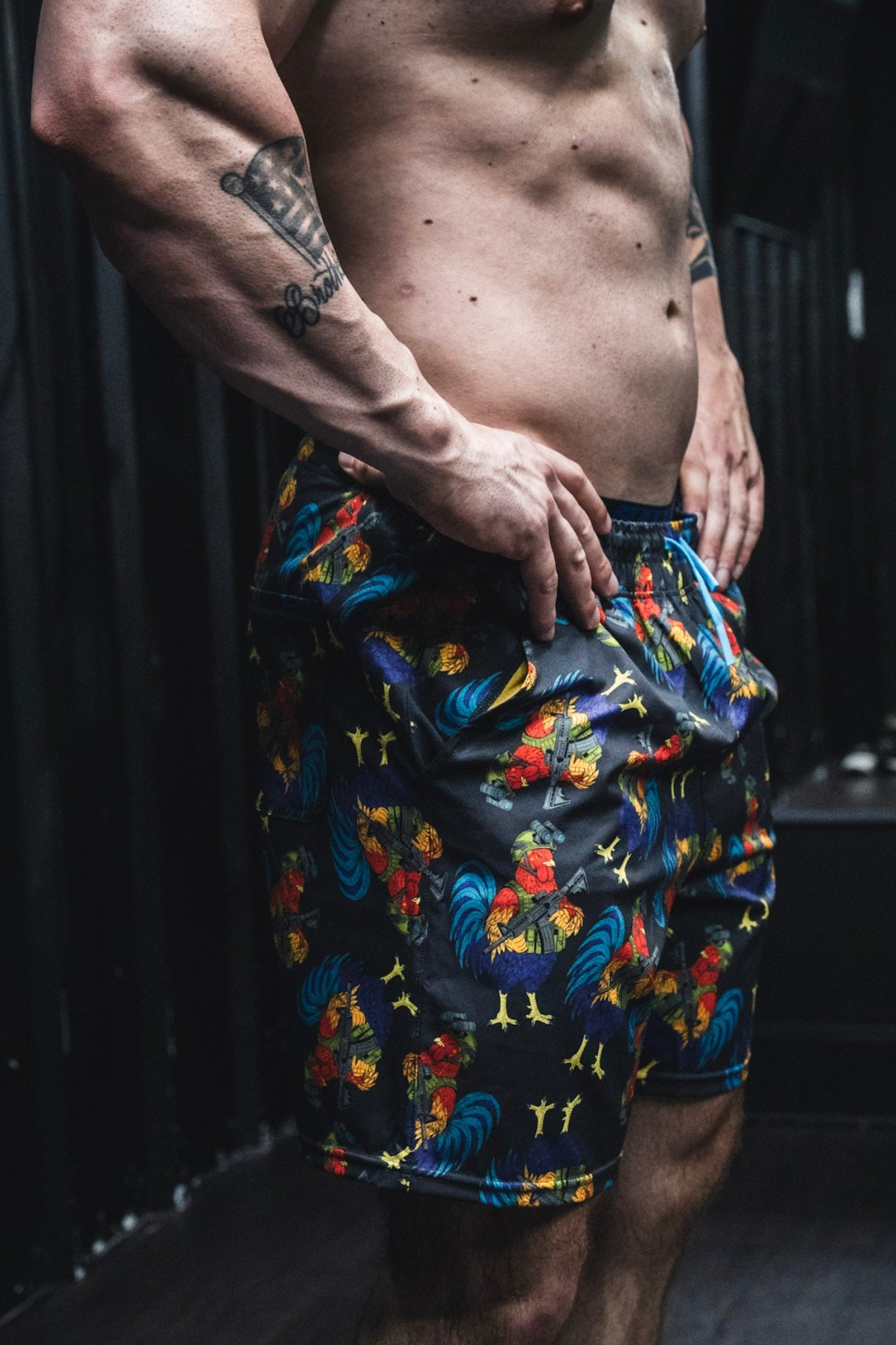 A person indoors wears Born United Private Label Men's Performance Hybrid Shorts 5.5" | Battle Bird, featuring rooster prints, hands on hips with a visible tattooed forearm and muscular torso. The dark background is out of focus.