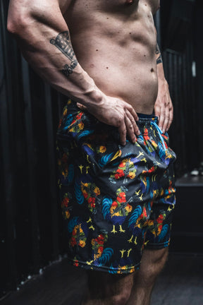 A person indoors wears Born United Private Label Men's Performance Hybrid Shorts 5.5" | Battle Bird, featuring rooster prints, hands on hips with a visible tattooed forearm and muscular torso. The dark background is out of focus.