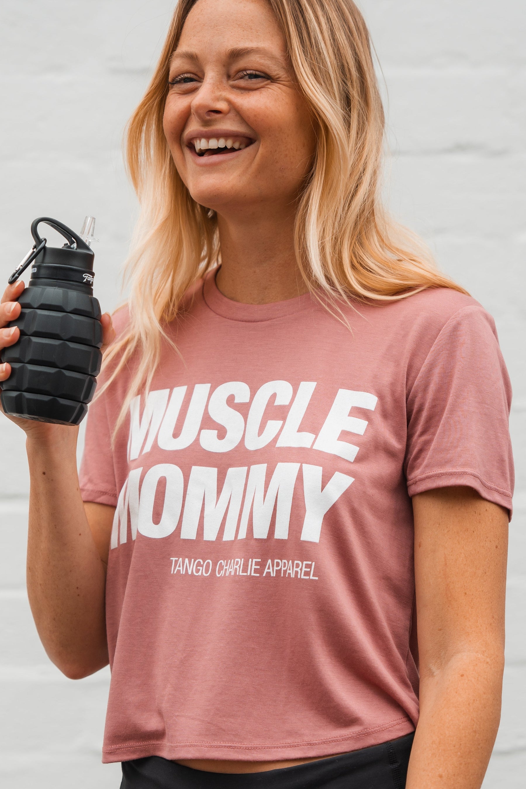 A fit, smiling woman with long blonde hair models the Muscle Mommy - Crop Tee by Tango Charlie Apparel, holding a black grenade-shaped water bottle. The simple background highlights her strength—she can deadlift 225 lbs.