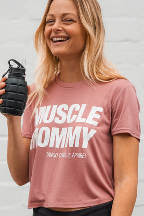 A fit, smiling woman with long blonde hair models the Muscle Mommy - Crop Tee by Tango Charlie Apparel, holding a black grenade-shaped water bottle. The simple background highlights her strength—she can deadlift 225 lbs.