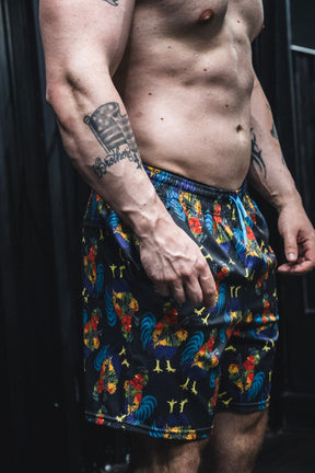 A shirtless, muscular person stands indoors, showing a tattooed arm and wearing Born United Private Label Men's Performance Hybrid Shorts 5.5" | Battle Bird, featuring colorful rooster designs and quick-drying fabric.