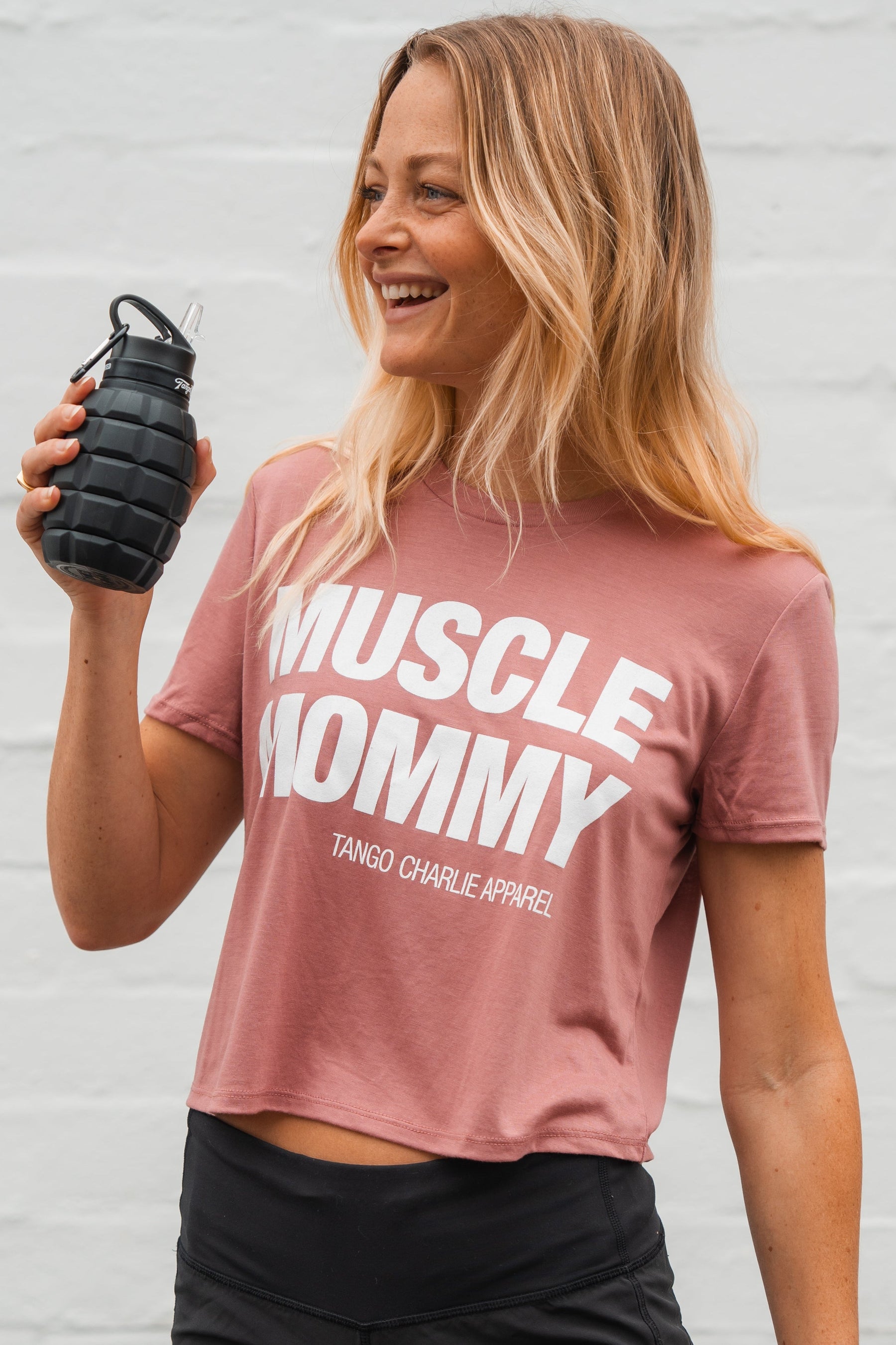 A fit woman with long blonde hair wears the Tango Charlie Apparel Muscle Mommy - Crop Tee in pink and holds a black, grenade-shaped water bottle, standing against a light background.