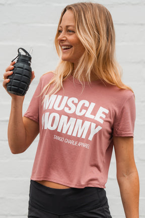 A fit woman with long blonde hair wears the Tango Charlie Apparel Muscle Mommy - Crop Tee in pink and holds a black, grenade-shaped water bottle, standing against a light background.