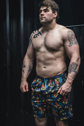 Man wearing colorful patterned shorts with tattoos on arms and chest against a dark background