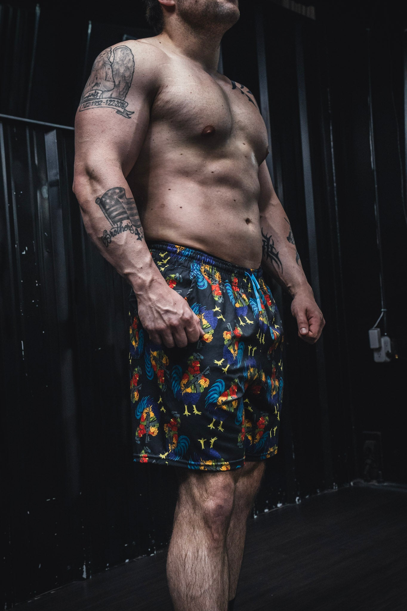 A muscular, tattooed man stands shirtless in Born United Private Label's Men's Performance Hybrid Shorts 5.5" | Battle Bird, featuring a rooster pattern, posing confidently in an industrial-style room.