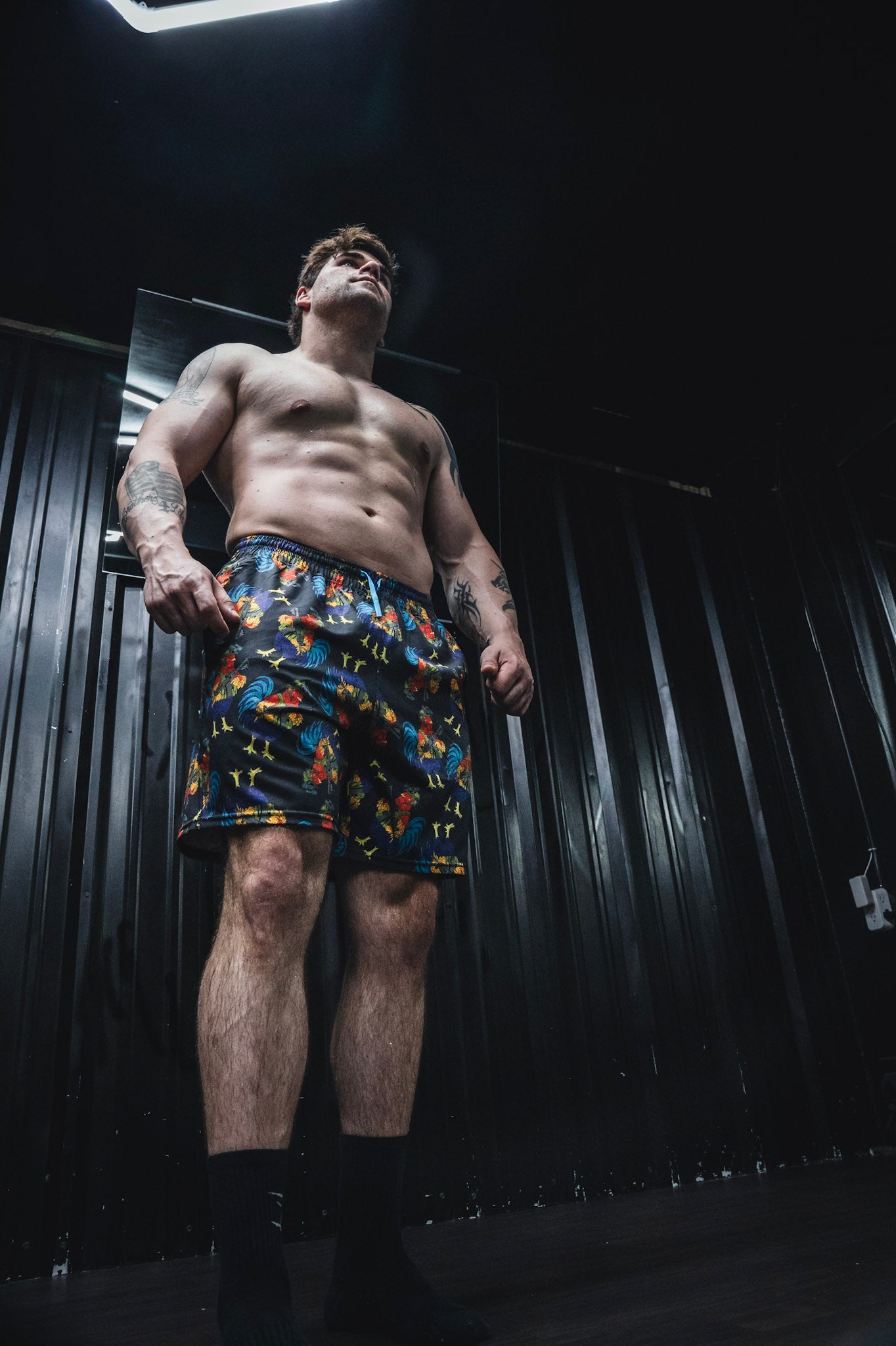 A shirtless man confidently stands in a dimly lit room with black walls, wearing Born United Private Label Men's Performance Hybrid Shorts 5.5" | Battle Bird, and looks upward.