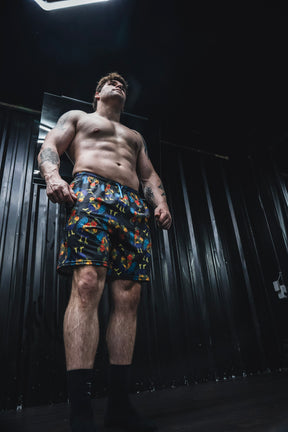 A shirtless man confidently stands in a dimly lit room with black walls, wearing Born United Private Label Men's Performance Hybrid Shorts 5.5" | Battle Bird, and looks upward.