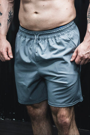 A person wearing Born United Private Label Men's Standard Performance Shorts | 5.5"- Gunmetal Gray stands with relaxed arms by their sides, upper body and head out of frame, and tattoos visible on their arms.