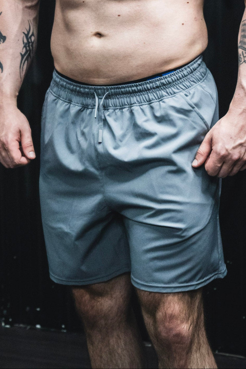 A person wearing Born United Private Label Men's Standard Performance Shorts | 5.5"- Gunmetal Gray stands with relaxed arms by their sides, upper body and head out of frame, and tattoos visible on their arms.