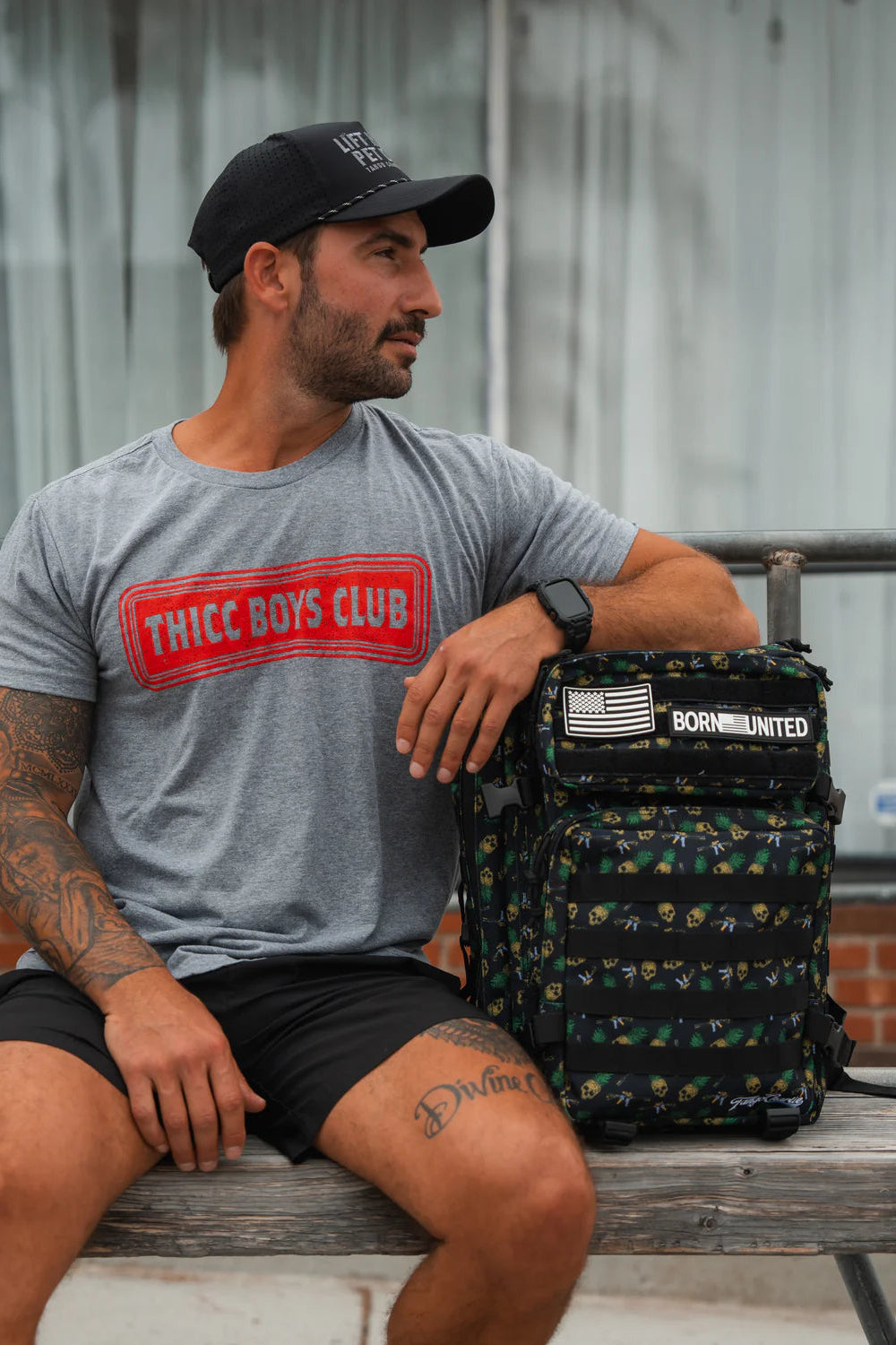 A bearded, tattooed man sits on a bench, wearing a black cap, gray Thicc Boys Club t-shirt, and black shorts. He leans on his versatile Born United Private Label TCA Tac-Pac - Tropic Thunder 2.0 - 45L (Warehouse), with patches like the U.S. flag. A large window is in the background.