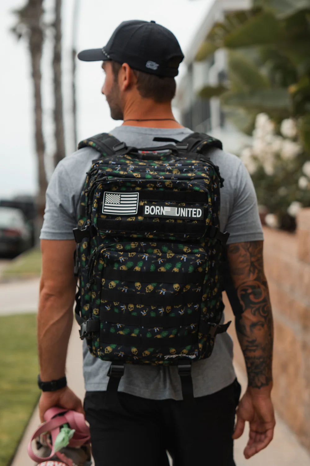A tattooed man strolls down the sidewalk in a black cap and gray t-shirt, carrying a TCA Tac-Pac - Tropic Thunder 2.0 (45L) from Born United Private Label, complete with an American flag and Born United patch. Pink and green items are in his left hand against trees and a wall backdrop.