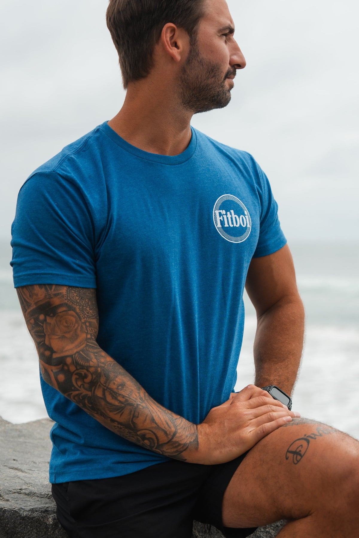 A bearded, tattooed man sits on a ledge by the ocean, wearing a blue Tango Charlie Apparel Pillsbury Fitboi T-Shirt made from soft ring-spun cotton and black shorts, gazing to the side.