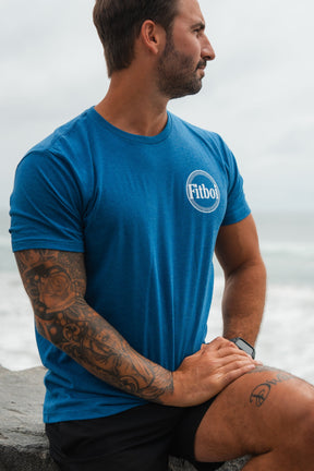 A bearded, tattooed man sits on a ledge by the ocean, wearing a blue Tango Charlie Apparel Pillsbury Fitboi T-Shirt made from soft ring-spun cotton and black shorts, gazing to the side.