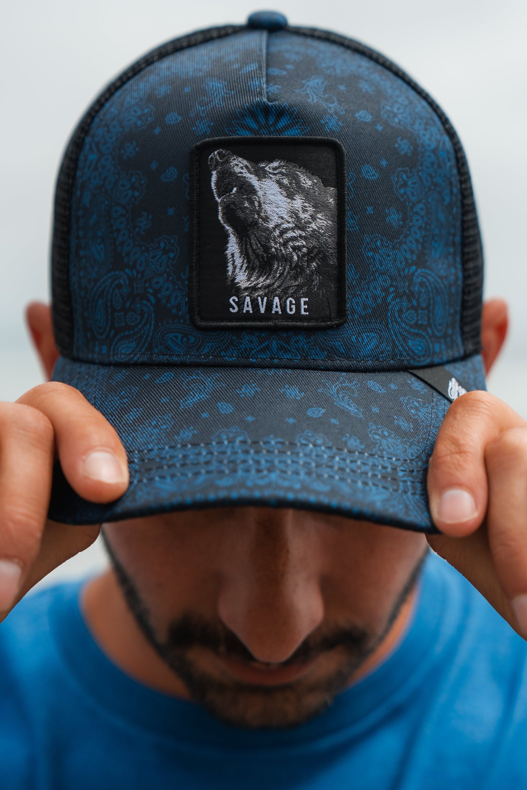 A man wearing a blue patterned trucker hat with a black mesh back and a square patch featuring a wolf illustration and the word 'SAVAGE'. He is holding the brim down over his eyes with both hands.