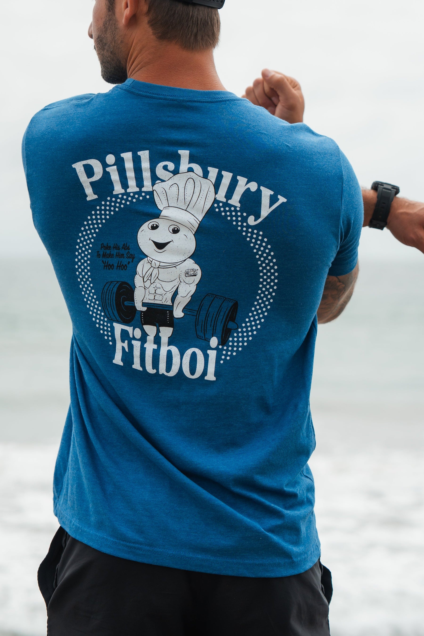 Person wearing a blue t-shirt with Pillsbury Fitboi graphic, standing by a body of water.