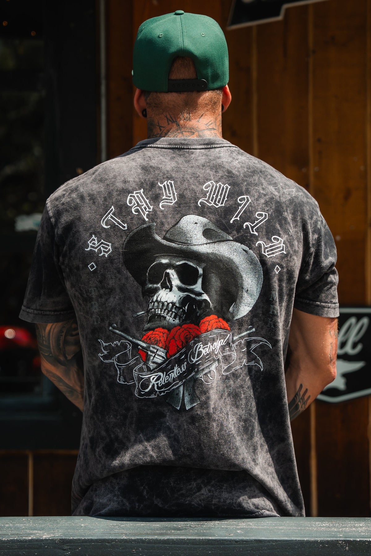 Someone wears the Stay Wild Premium T-Shirt - Stone by Relentless Betrayal, featuring tie-dye colors and a bold back graphic with a skull in a cowboy hat, red roses, and STAY WILD above Relentless Bandit, paired with a green cap.
