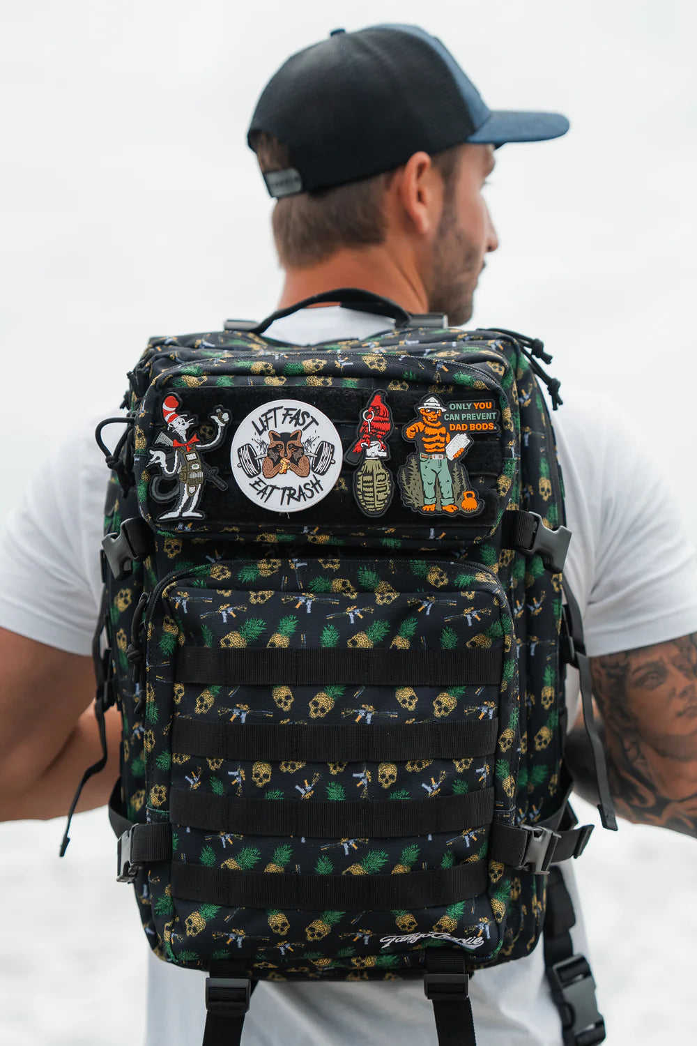 A man in a black cap and white t-shirt carries the TCA Tac-Pac Tropic Thunder 2.0 backpack by Born United Private Label, featuring colorful skull and beach-themed patches for tactical versatility, against a blurred sand and ocean backdrop.