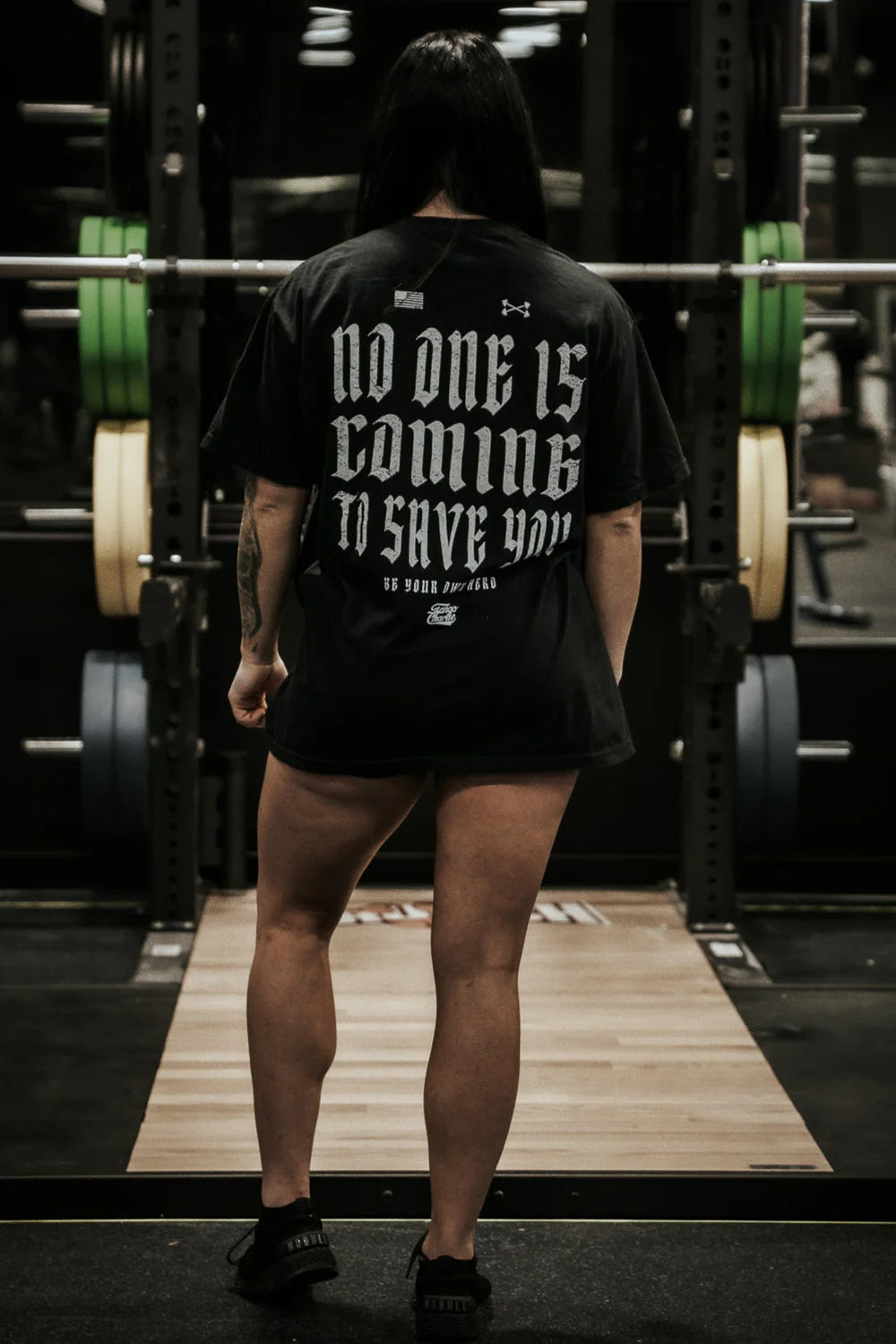 A person with long dark hair stands in front of a squat rack, wearing the Tango Charlie Apparel "No One Is Coming To Save You Tee - Black," featuring the bold motivational quote on the back.