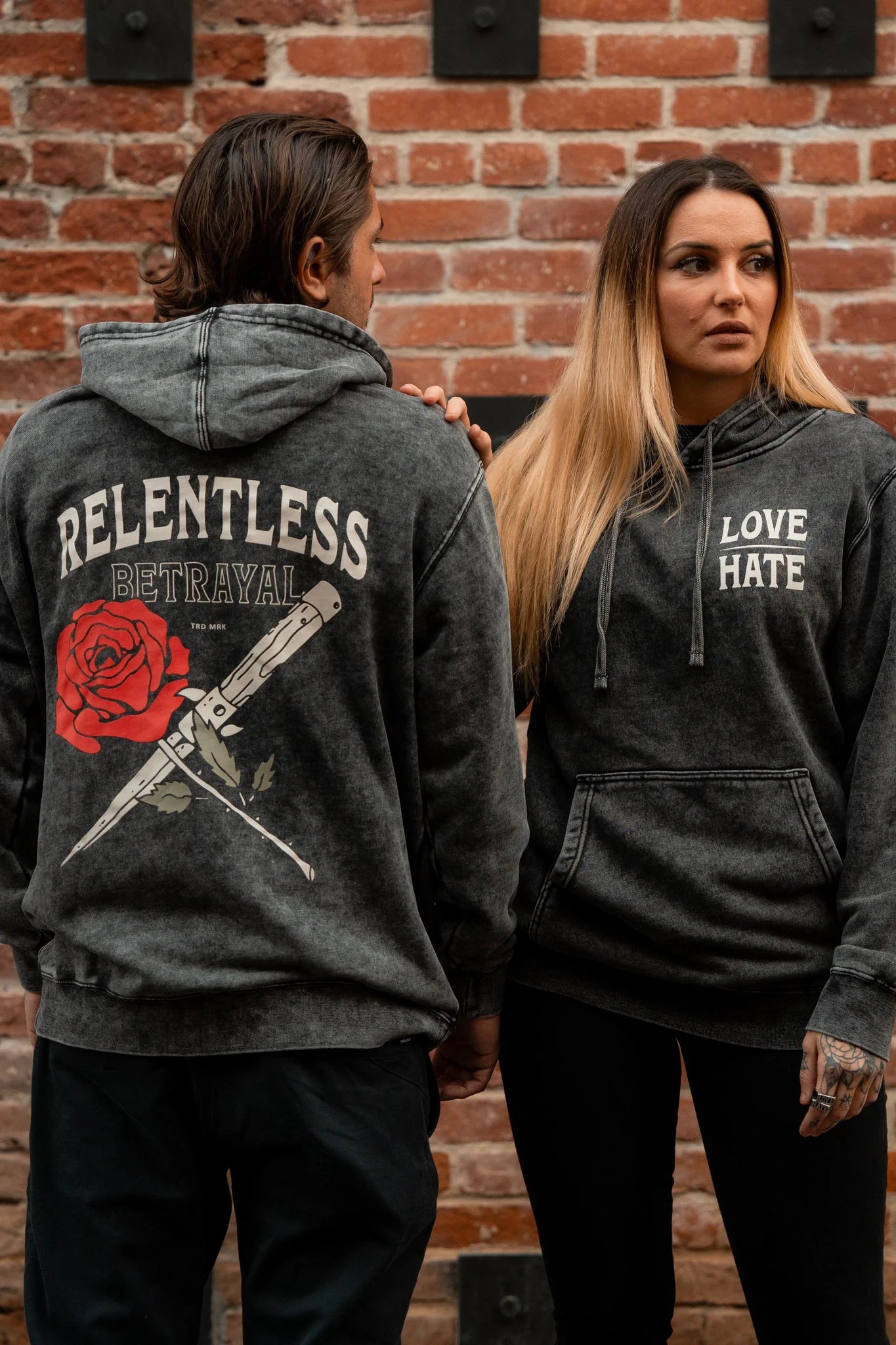 Two people in dark hoodies stand by a brick wall. The one on the left showcases Relentless Betrayals rose and knife design, while on the right, the Love/Hate Premium Hoodie from our unisex line flaunts bold text on a cotton/polyester blend.