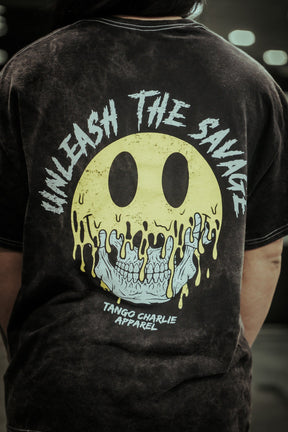 A person wears the Unleash The Savage Premium Tee by Tango Charlie Apparel, featuring a large back graphic: a melting yellow smiley revealing a skull, with "UNLEASH THE SAVAGE" above and the brand name below.