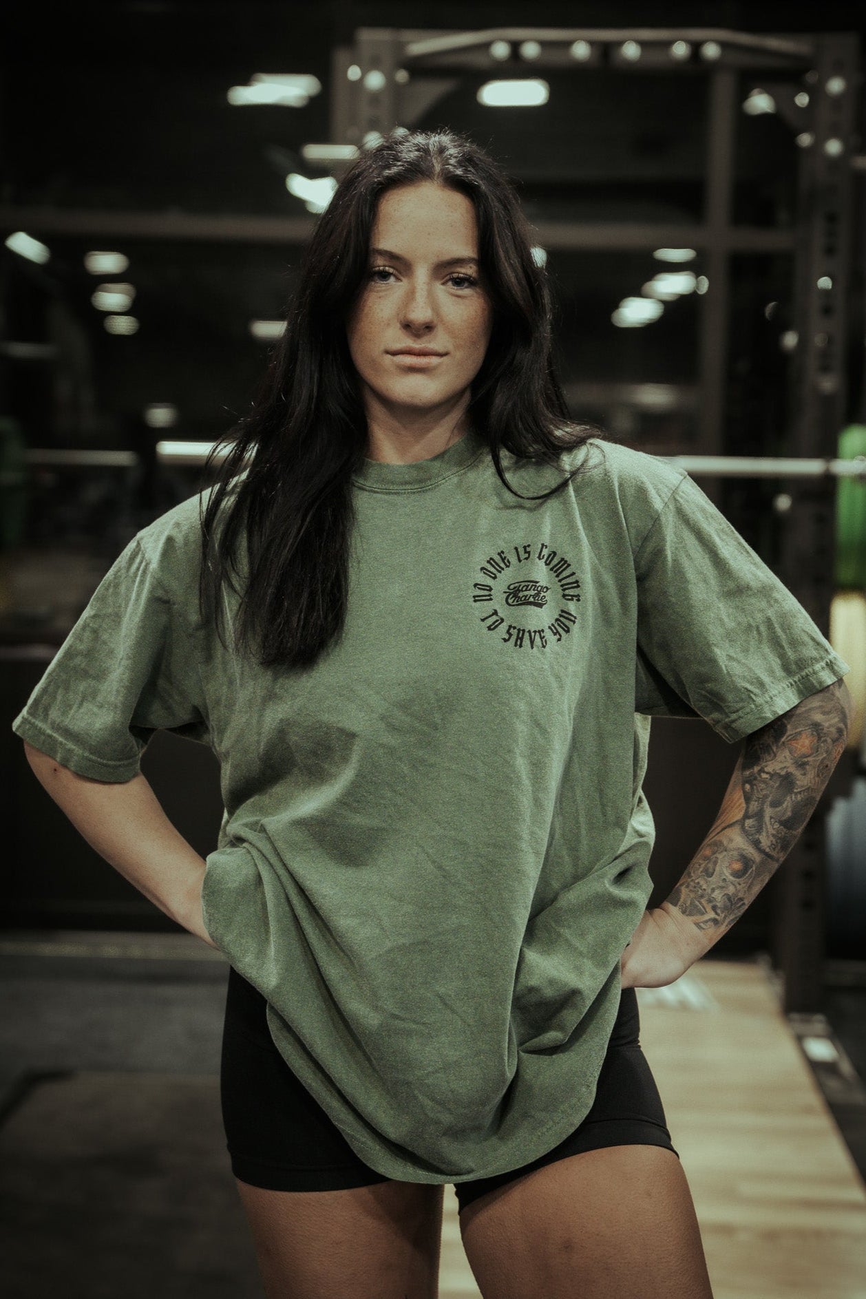 A woman with long dark hair stands confidently in a gym, hands on hips, showing her forearm tattoo. She wears Tango Charlie Apparel’s No One Is Coming To Save You Hemp Premium Tee in green with black shorts, exuding strength.