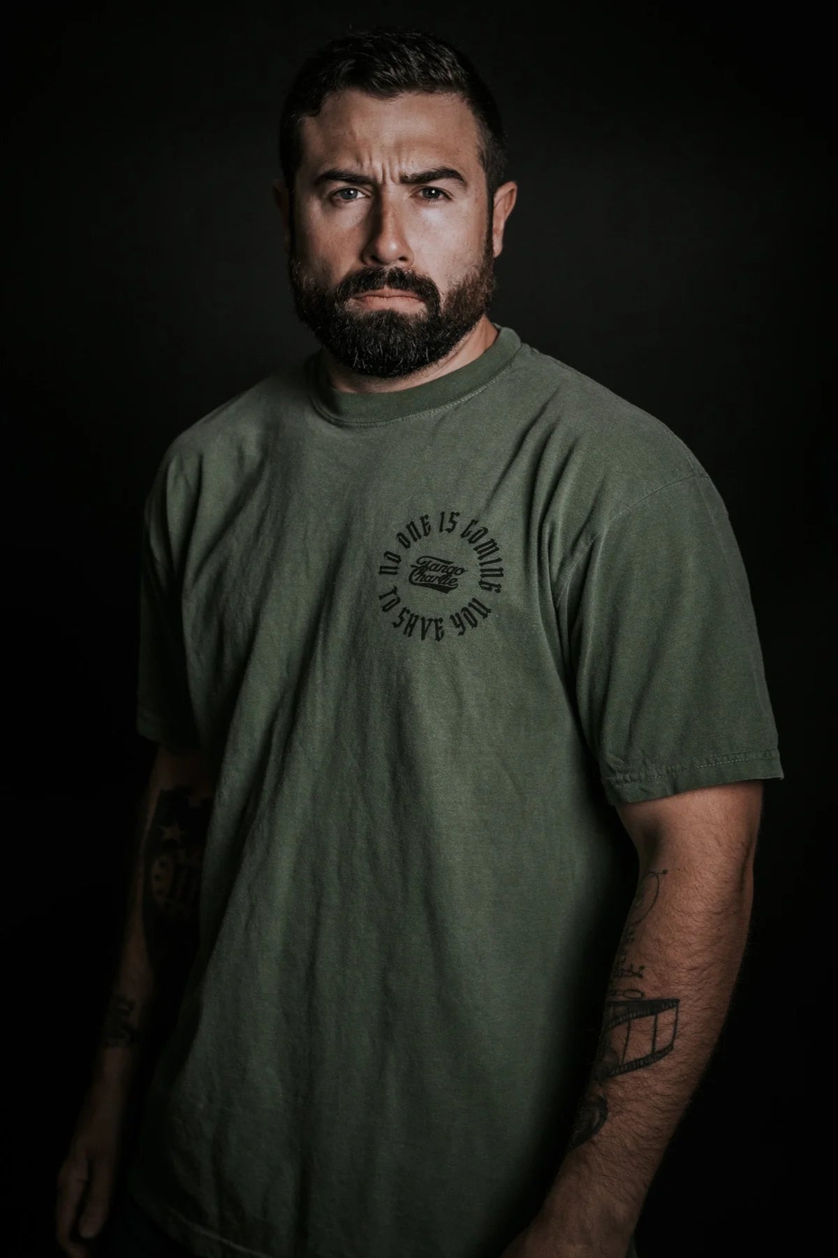A man with a serious expression, full beard, and short dark hair stands against a dark background, wearing a loose-fitting olive green "No One Is Coming To Save You Tee- Hemp" by Tango Charlie Apparel, with tattoos on his forearms.