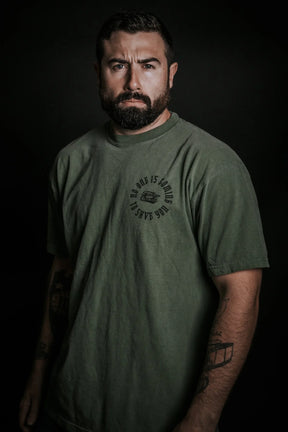 A man with a serious expression, full beard, and short dark hair stands against a dark background, wearing a loose-fitting olive green "No One Is Coming To Save You Tee- Hemp" by Tango Charlie Apparel, with tattoos on his forearms.