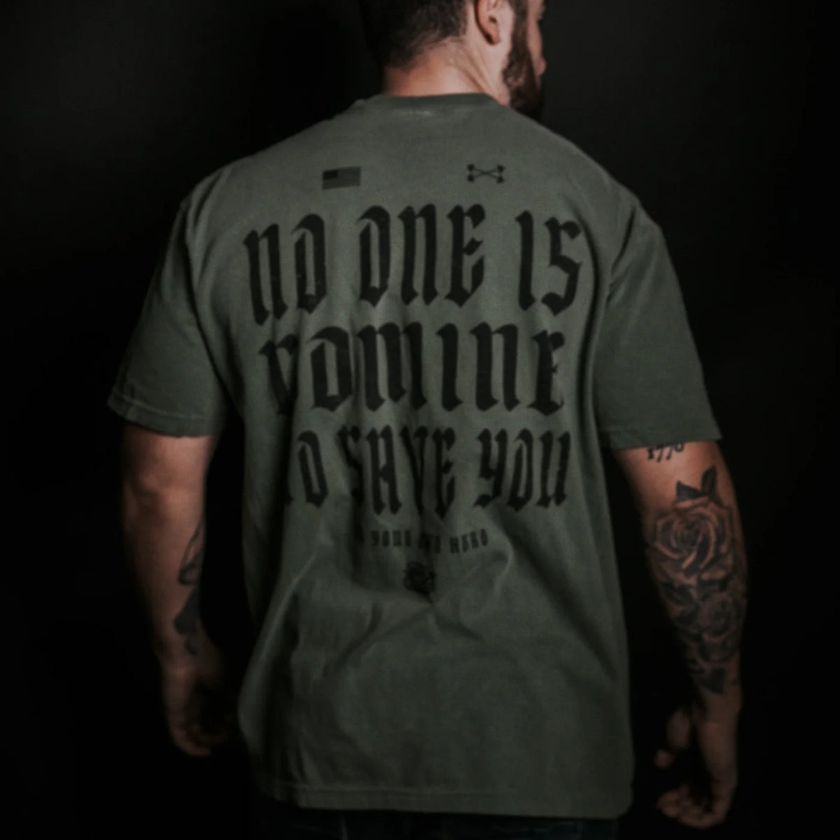 A man with tattooed arms, facing away, wears the Tango Charlie Apparel No One Is Coming To Save You Tee-Hemp in olive green with bold black text, set against a dark background.