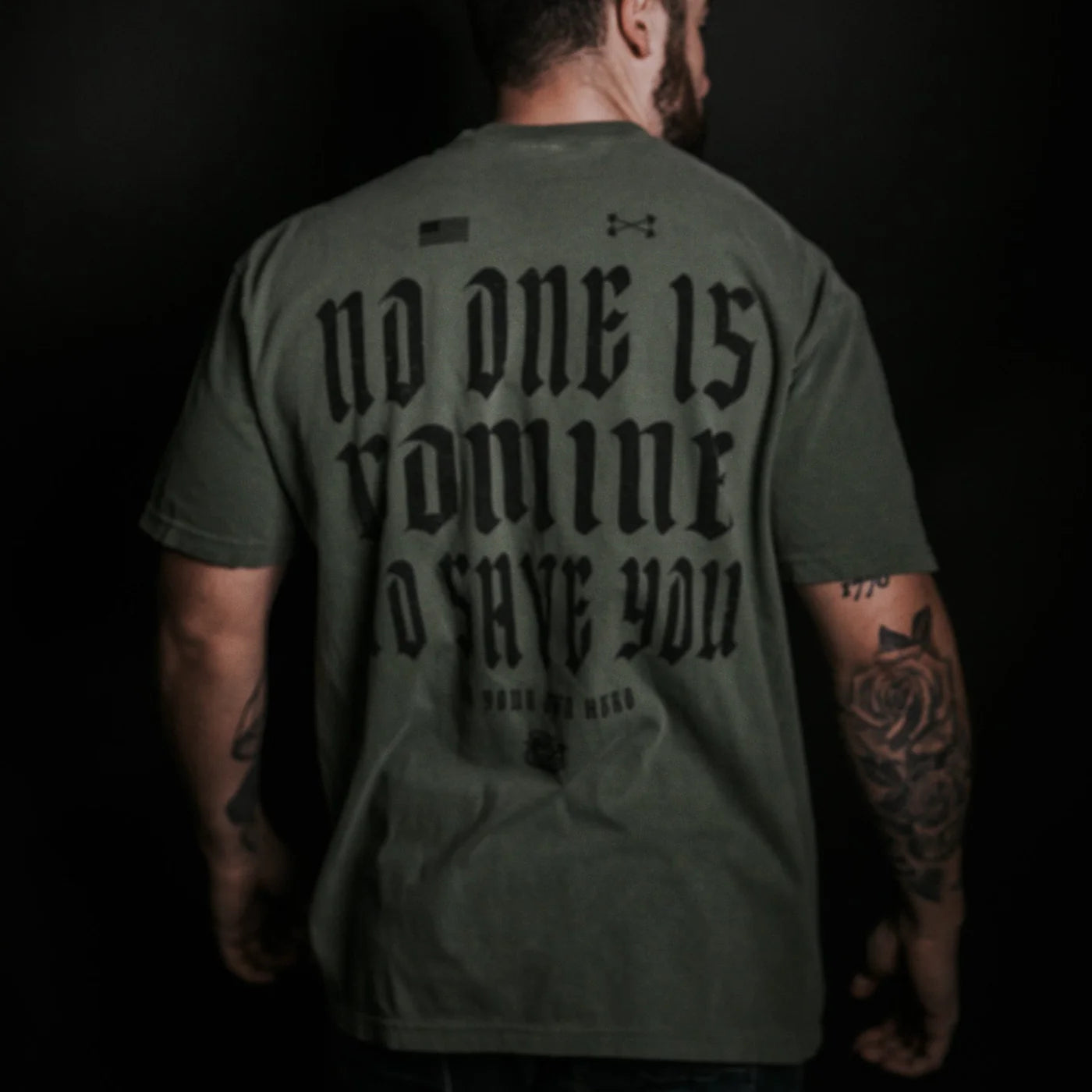 A man with tattooed arms, facing away, wears the Tango Charlie Apparel No One Is Coming To Save You Tee-Hemp in olive green with bold black text, set against a dark background.