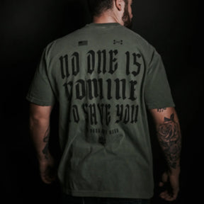 A man with tattooed arms, facing away, wears the Tango Charlie Apparel No One Is Coming To Save You Tee-Hemp in olive green with bold black text, set against a dark background.