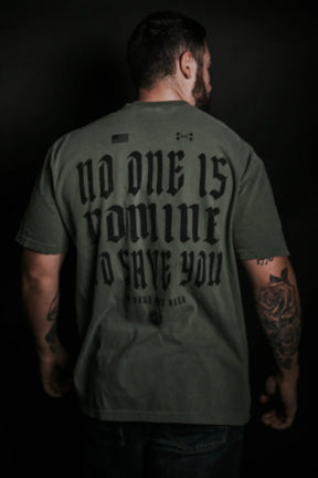 A tattooed man faces away from the camera, wearing Tango Charlie Apparel’s No One Is Coming To Save You Tee—Hemp, featuring the bold gothic message “NO ONE IS COMING TO SAVE YOU—GET UP” on a dark green shirt against a dark background.