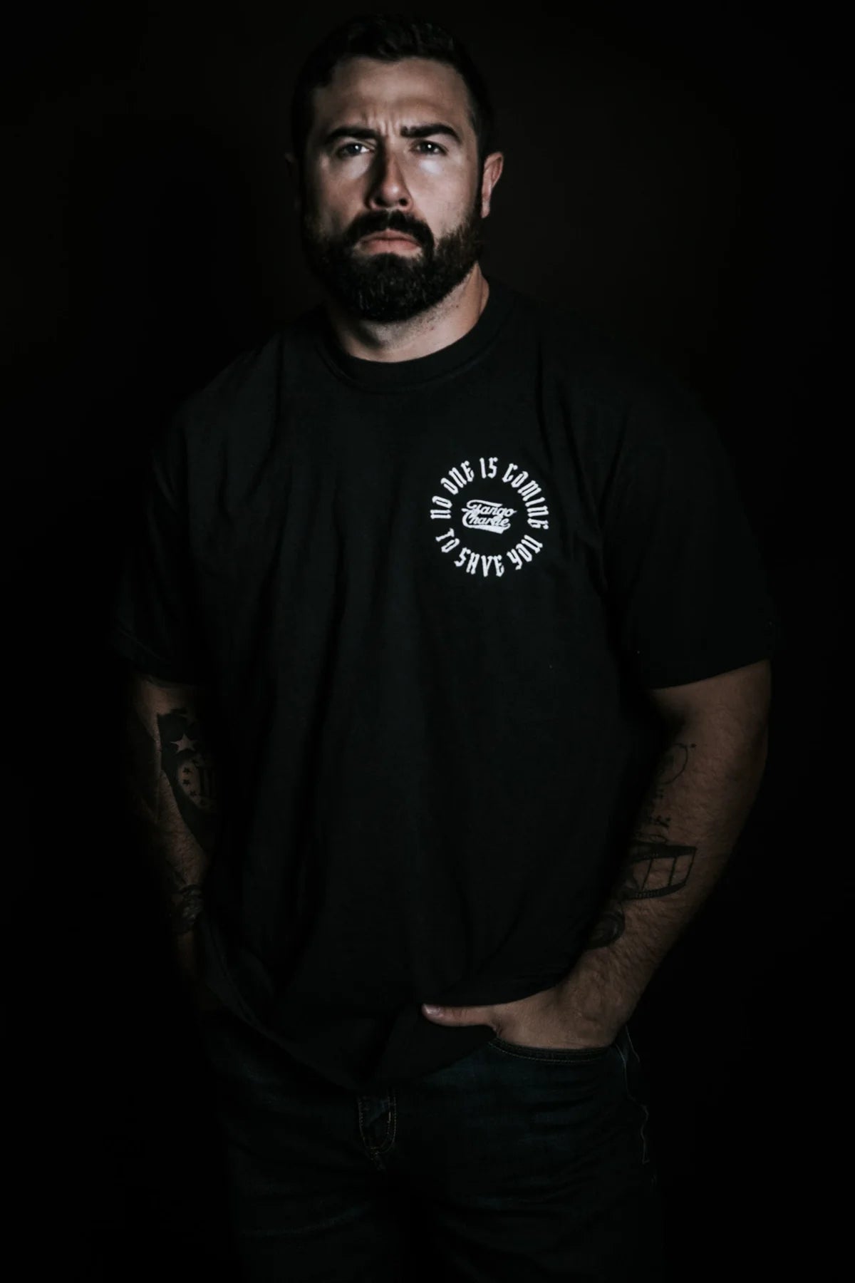 A bearded man with visible tattoos stands against a dark background, hands in his jeans pockets, wearing the "No One Is Coming To Save You Tee - Black" by Tango Charlie Apparel.