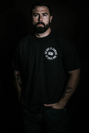 A bearded man with visible tattoos stands against a dark background, hands in his jeans pockets, wearing the "No One Is Coming To Save You Tee - Black" by Tango Charlie Apparel.