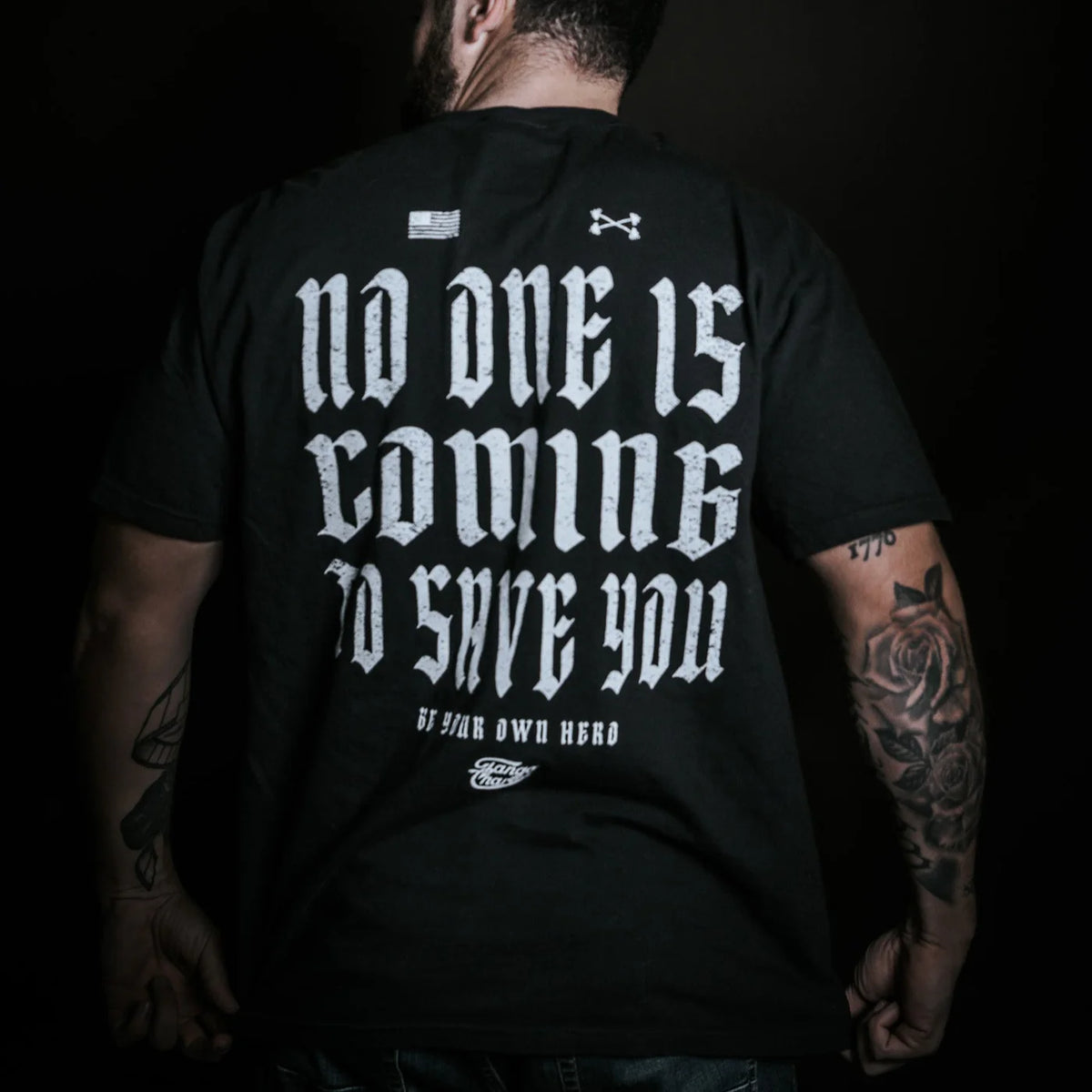 Wearing the No One Is Coming To Save You Tee in black by Tango Charlie Apparel, a tattooed individual stands confidently against a dark backdrop—boldly displaying the quote, "No One Is Coming To Save You. Be your own hero.