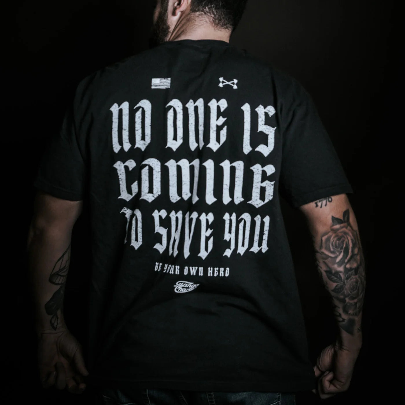 Wearing the No One Is Coming To Save You Tee in black by Tango Charlie Apparel, a tattooed individual stands confidently against a dark backdrop—boldly displaying the quote, "No One Is Coming To Save You. Be your own hero.