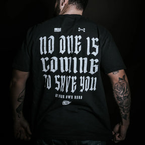 Wearing the No One Is Coming To Save You Tee in black by Tango Charlie Apparel, a tattooed individual stands confidently against a dark backdrop—boldly displaying the quote, "No One Is Coming To Save You. Be your own hero.